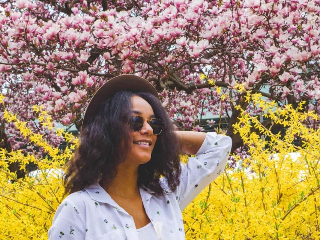 woman wearing sunglasses holding hat among yellow flowers and pink cherry blossoms