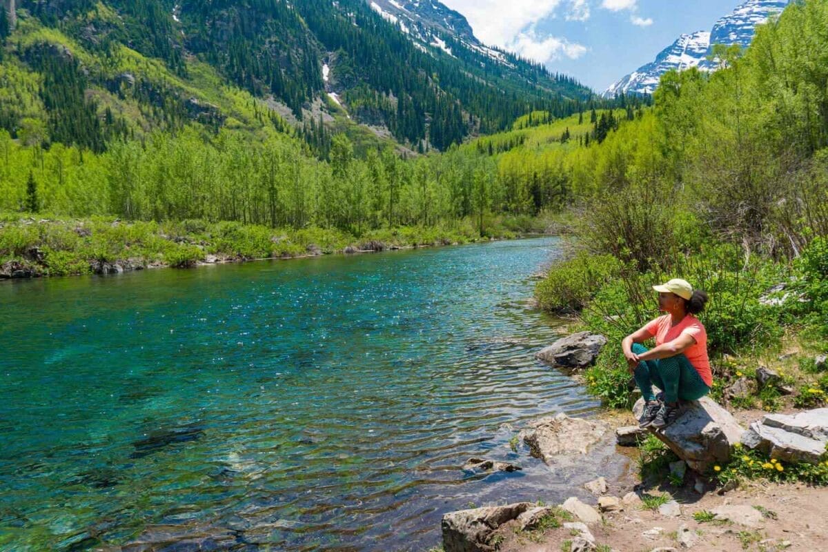 black female hiker sitting on a rock by a clear blue stream river and surrounded by mountains and trees