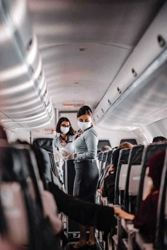 two flight attendants wearing masks while walking down the plane aisle with food and beverage service with food cart