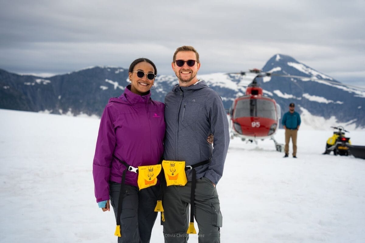 travel couple posing in front of a helicopter in the snow and surrounded by mountains