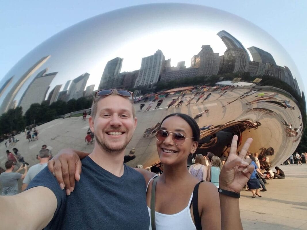Travel couple standing in front of a silver bean shaped sculpture, with a city skyline in the reflection