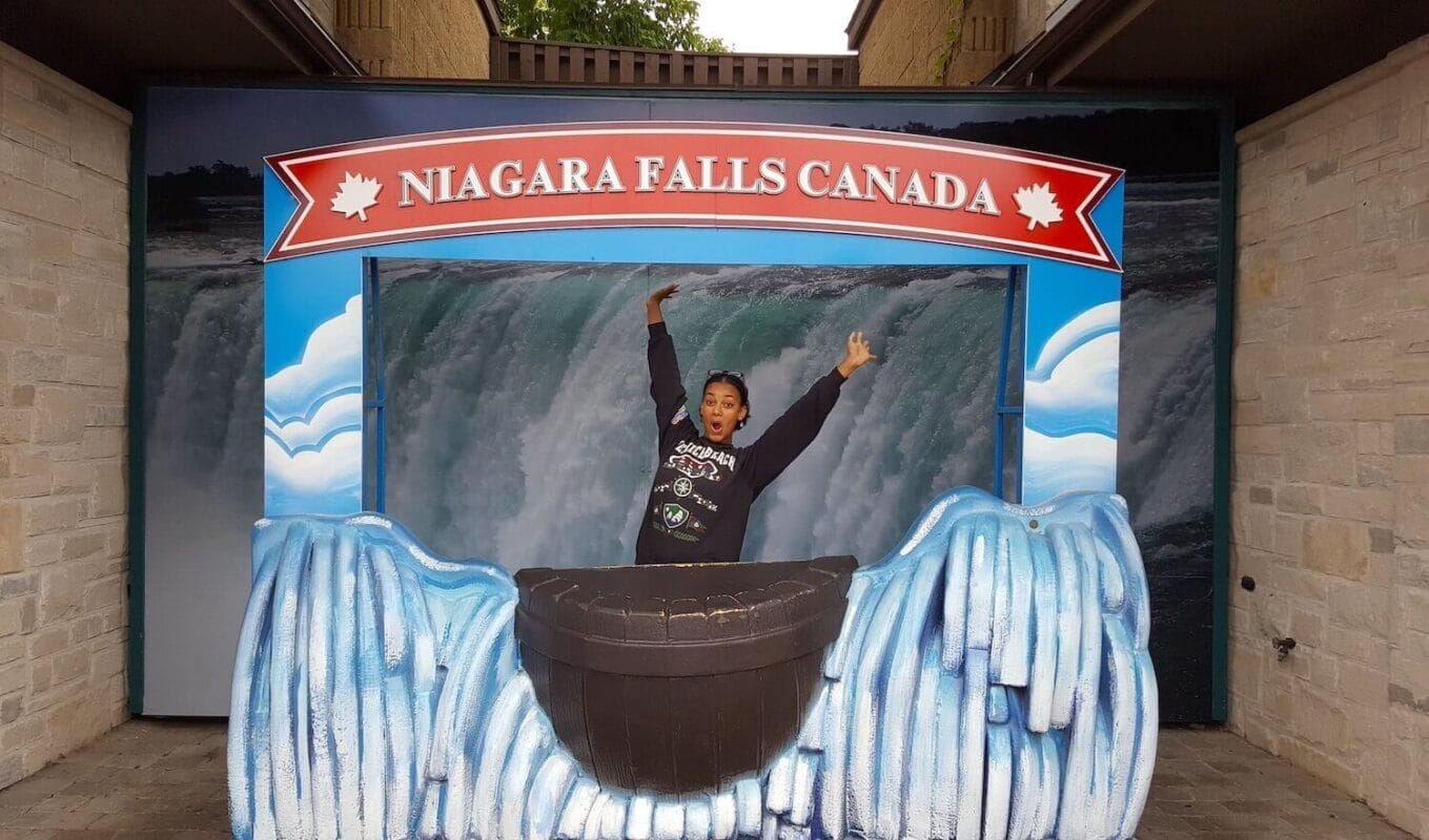 Tourist posing in a cardboard photo prop that says Niagara Falls Canada and has a mockup of a barrel falling down painted waterfalls