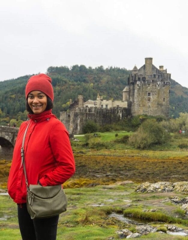 Afro latina traveler wearing a red beanie and windbreaker and standing in front of a castle and lush mossy ground