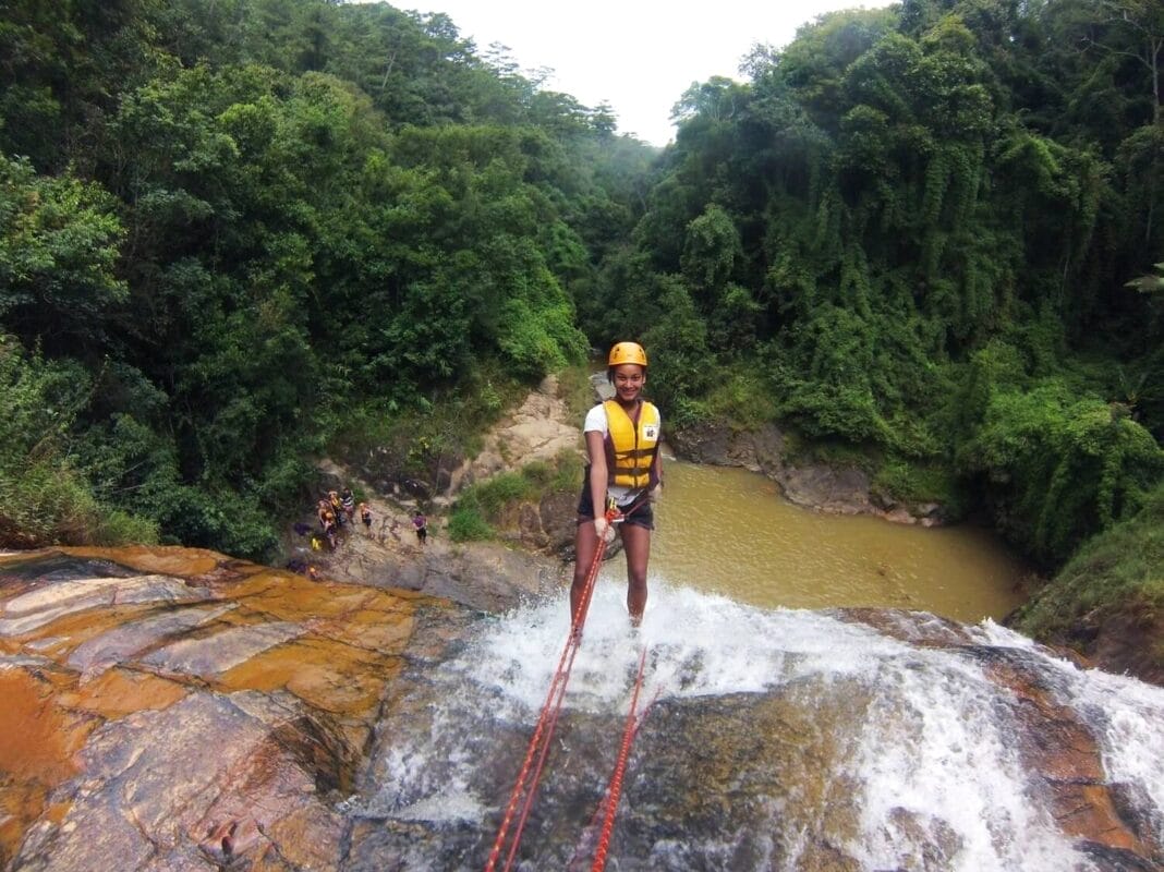 Black outdoor traveler rappelling down a waterfall, smiling up at the camera