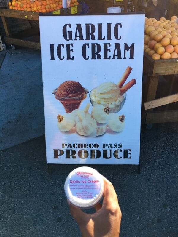 holding a cup of ice cream in front of a sign that says Garlic Ice Cream Pacheco Pass