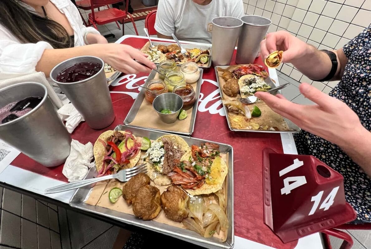 four people sitting at a table with platters of tacos and potatoes